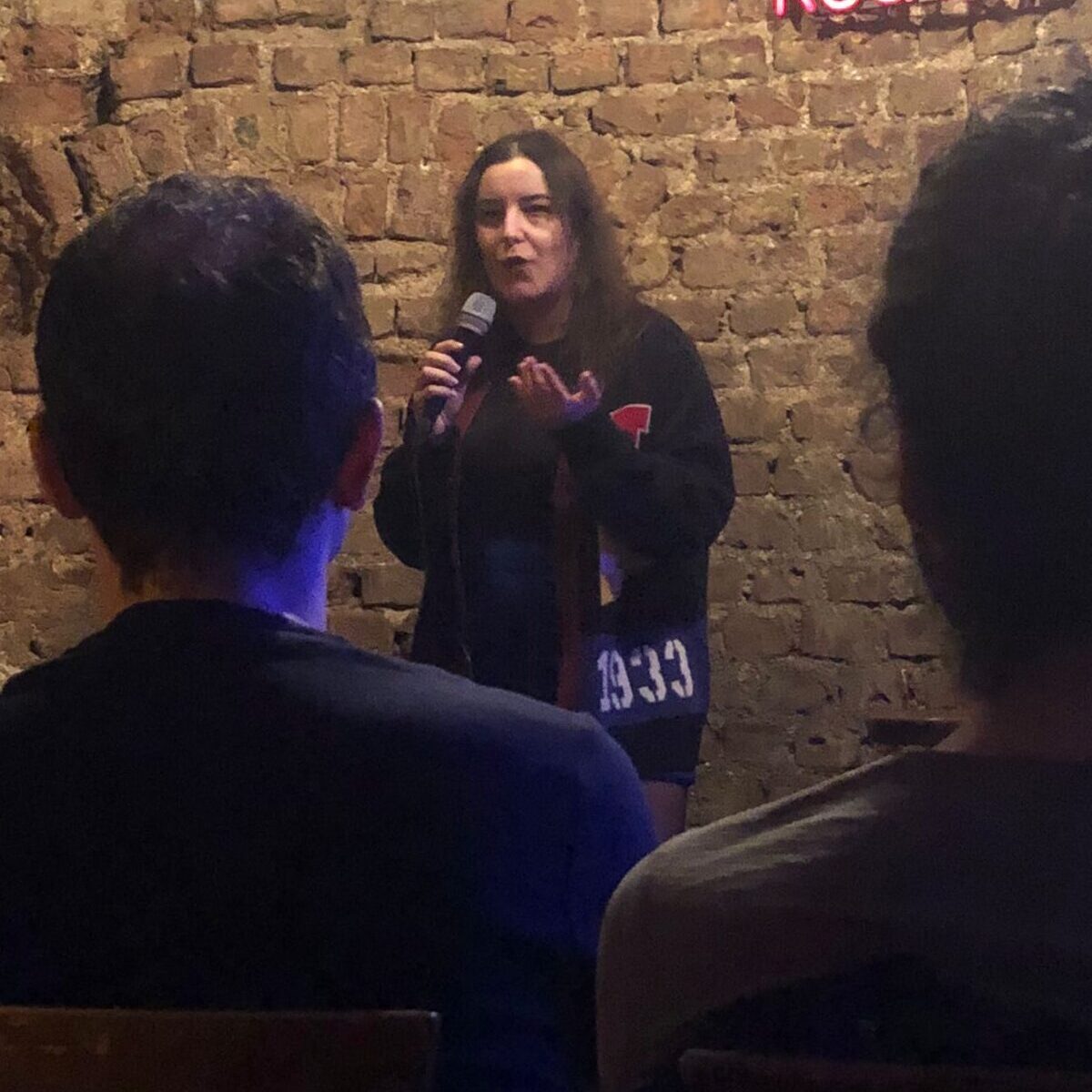 A photo of Cansu Pylkkänen performing stand-up comedy on stage, engaging the audience with humor and energy. The setting is intimate and lively, reflecting the dynamic atmosphere of the show.